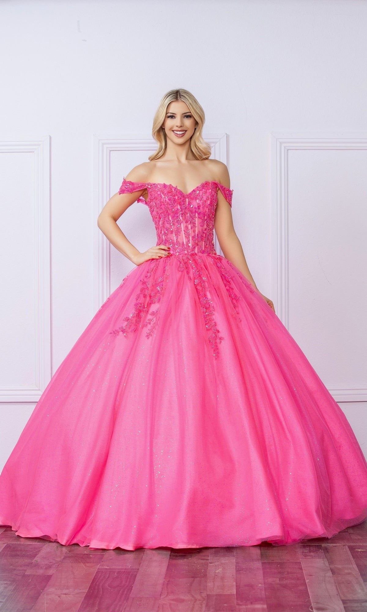 A woman stands indoors wearing the Bright Off-the-Shoulder Long Prom Ball Gown H1349, featuring a full tulle skirt and floral embellishments—a stunning choice for any formal event.